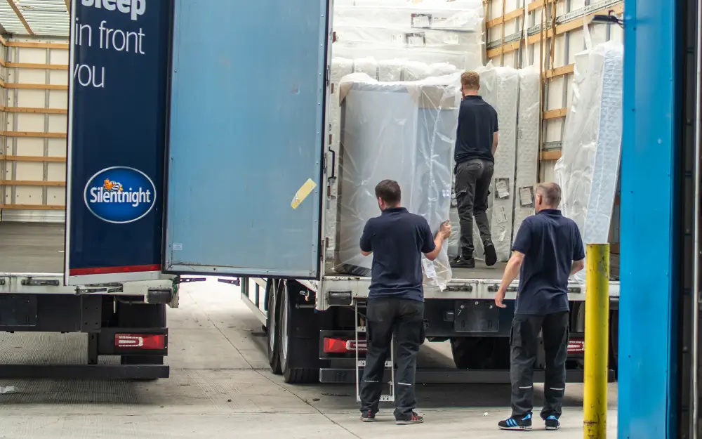 Three delivery workers loading large, plastic-wrapped mattresses into the back of a Silentnight branded delivery truck at a warehouse loading bay.