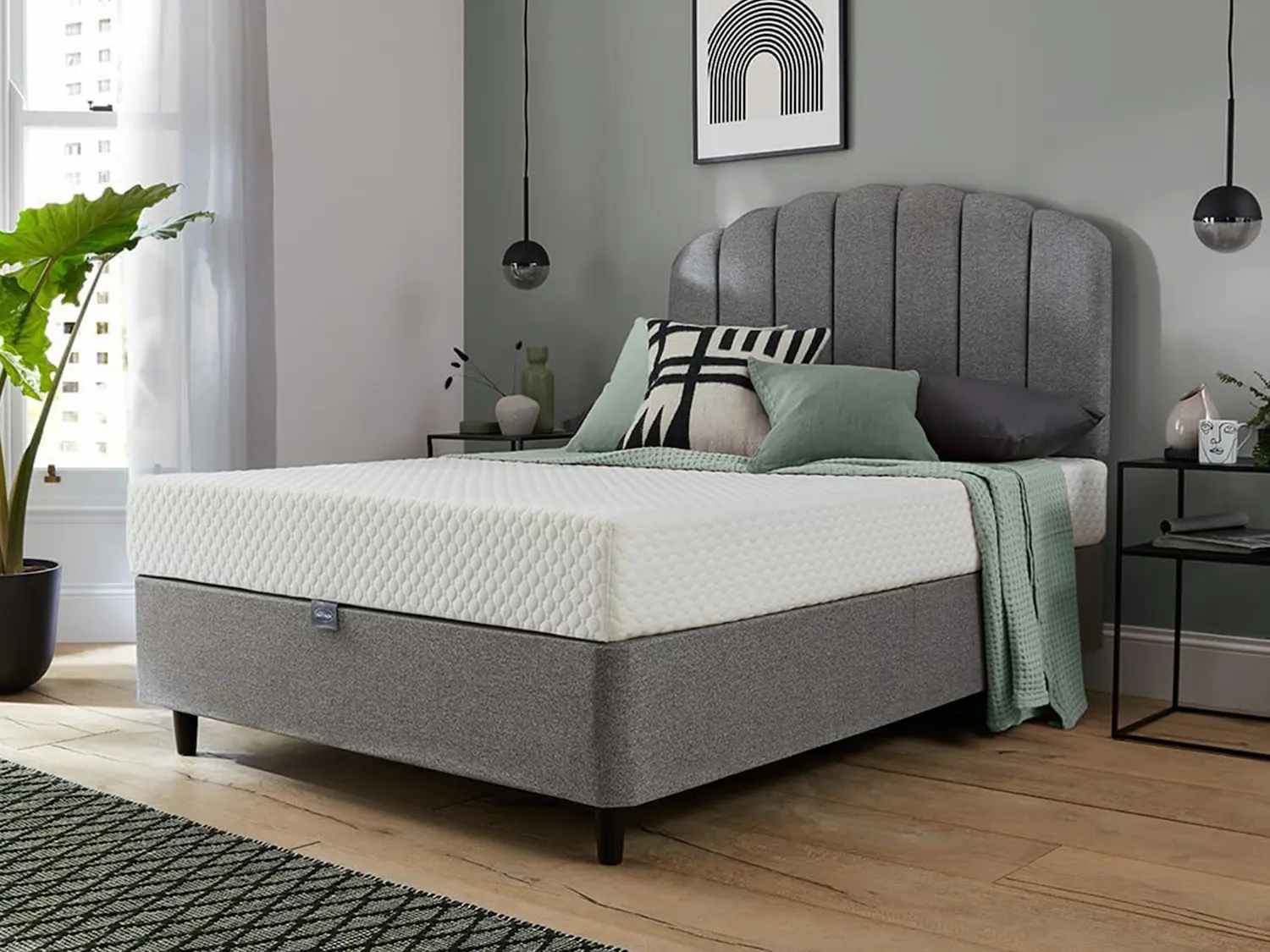 Silentnight 7 Zone Memory Foam Rolled Mattress in white quilted cover, displayed on a grey upholstered divan base with a scalloped headboard in a styled bedroom with green accents and wooden flooring.