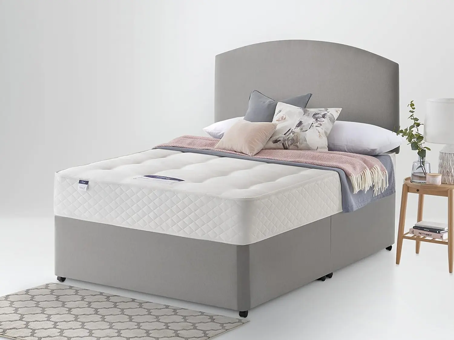 Silentnight Miracoil Ortho Mattress with white diamond-quilted cover, shown on a grey divan bed base with a curved upholstered headboard, styled with pink and grey bedding against a light grey background.