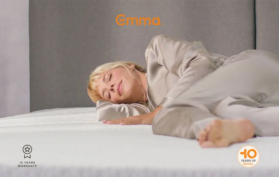A woman sleeping comfortably on her side on a white Emma mattress, wearing silk pyjamas. The image includes the Emma logo and icons for a 10-year warranty and a '10 Years of Emma' anniversary seal.
